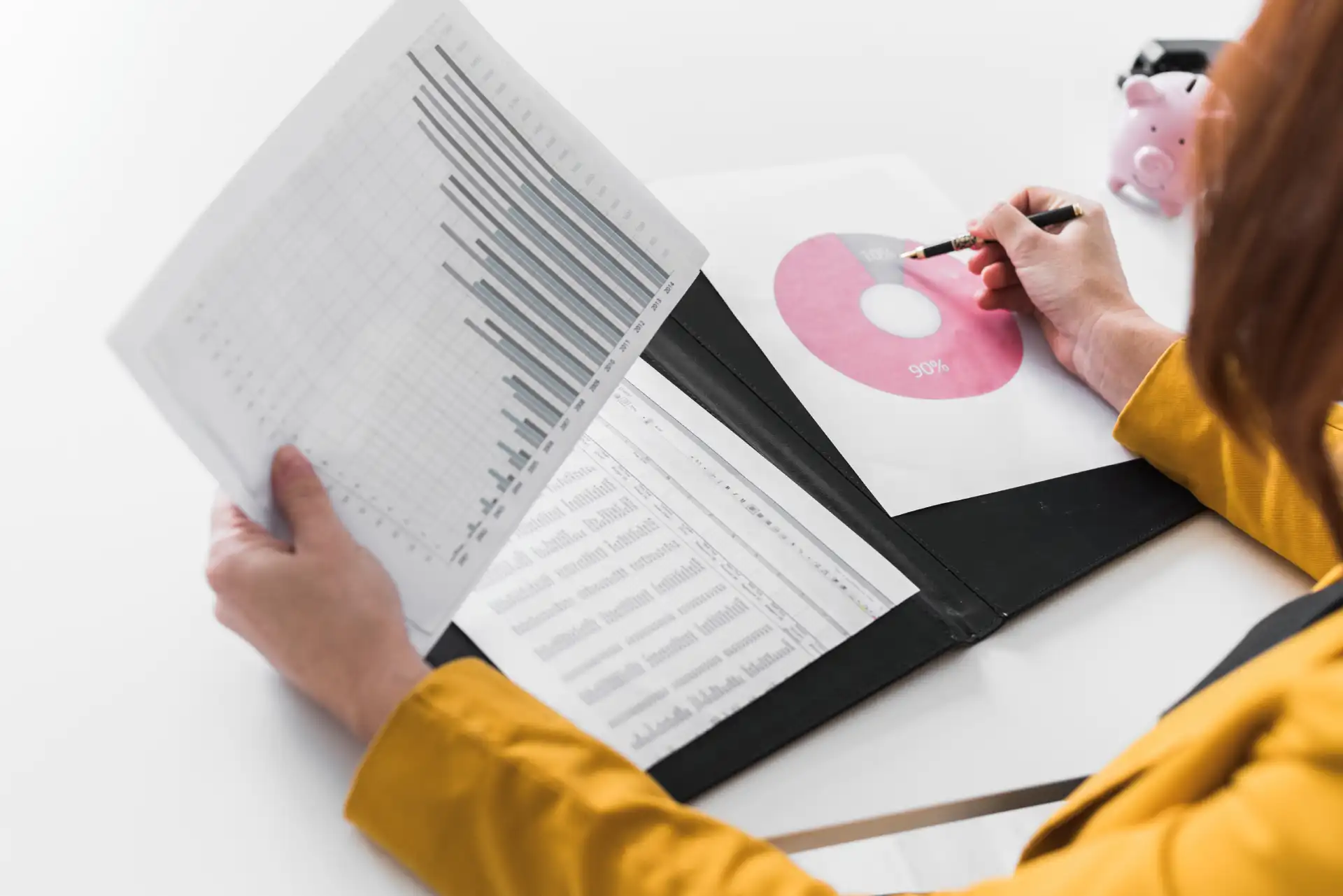 Reviewing 90% charts in a black folder; person in yellow blazer analyzing data with a pink piggy bank in the background