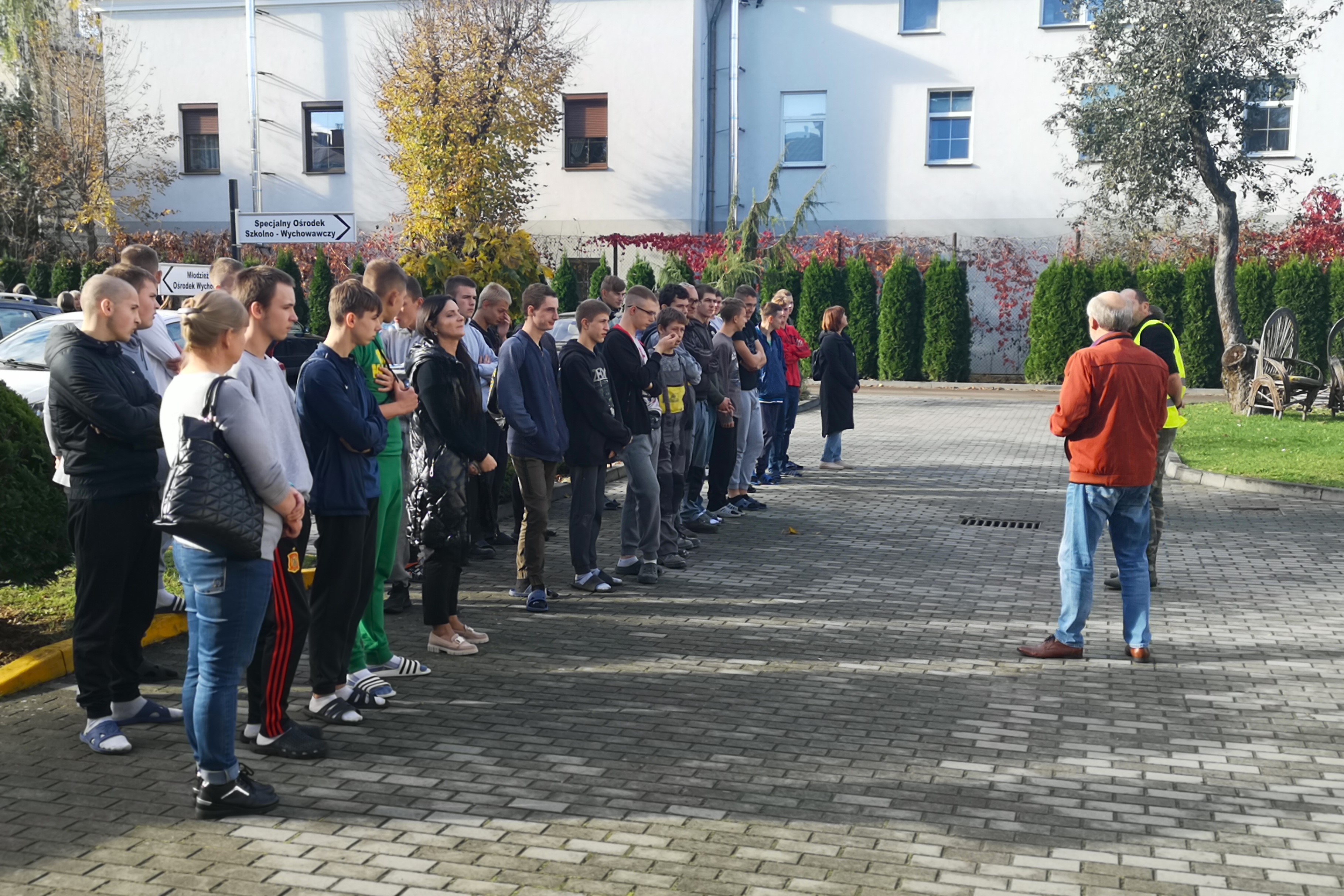 Próbna ewakuacja przeciwpożarowa | Chłopcy stojący w dwuszeregu i ich nauczyciele na placu szkoły - widok z boku. Przed nimi instruktor BHP w rudej koszuli i niebieskich dżinsach. W tle zieleń drzew, żywopłotu oraz zabudowania..jpg