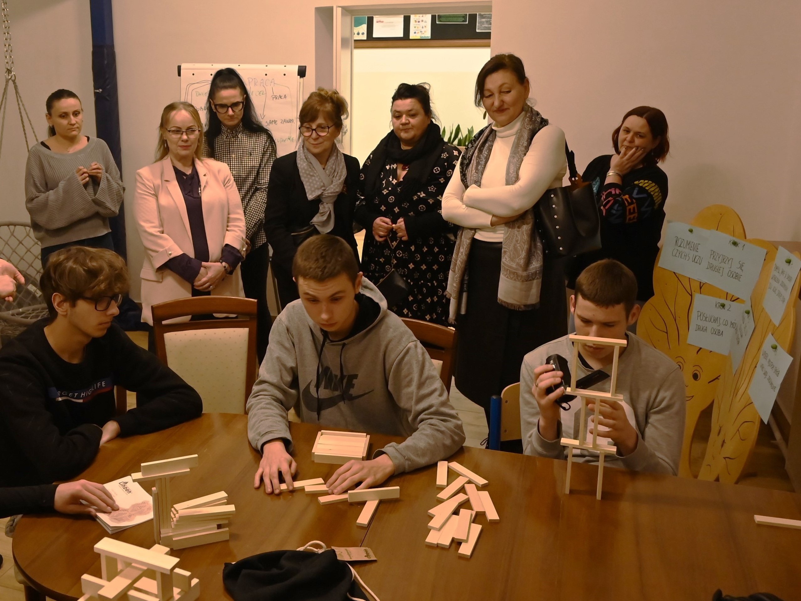 Zakończenie projektu Fajna Ferajna | Trzech chłopców siedzi przy stole układając drewniane kloci. Za nimi siedem dorosłych kobiet przypatruje się zajęciom.JPG