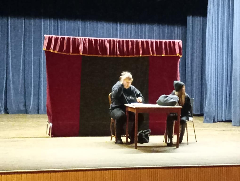 Spektakl profilaktyczny | Scena teatru, na środku stoi stolik i dwa krzesłą, na nich siedzą dwie dziewczyny unrane na czarno. Na stole plecak szkolny. Z tyłu parawan.jpg