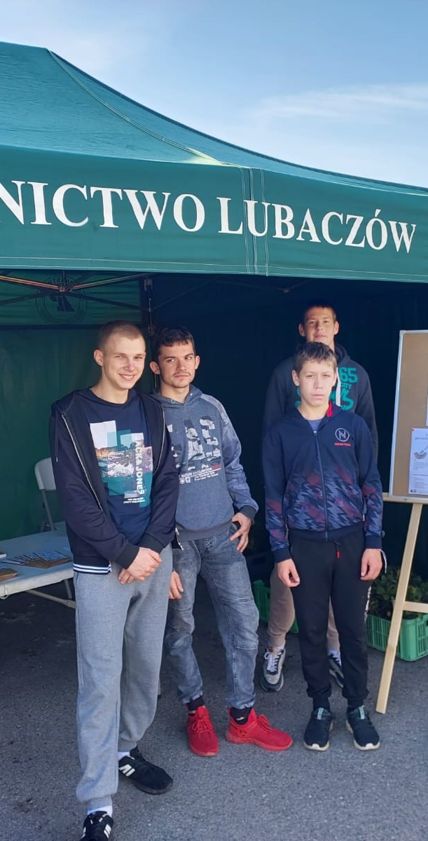 Sadzimy z Prezydentem RP | Czterechwychowanków MOW stoi przed zielonym namiotem Nadleśnictwa Lubaczów, za nimi pod namiotem stół z materiałami reklamowymi, obok fragment sztalugi z plakatem..jpg