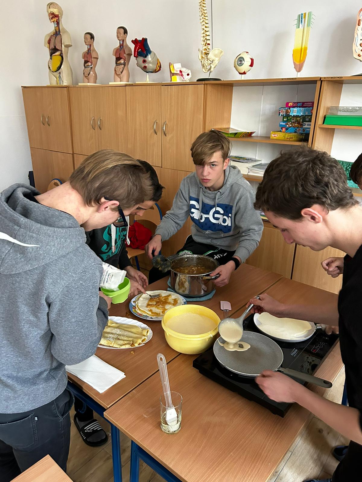 Andrzejkowe wróżby i nie tylko. | Czterech chłopców stoi wokół dwóch zsuniętych razem stolików szkolnych. Na blatach elektryczna kuchenka, na której smażą się naleśniki, obok miska z ciastem, talerze z gotowymi plackami, które chłopcy smarują dżemem..jpg