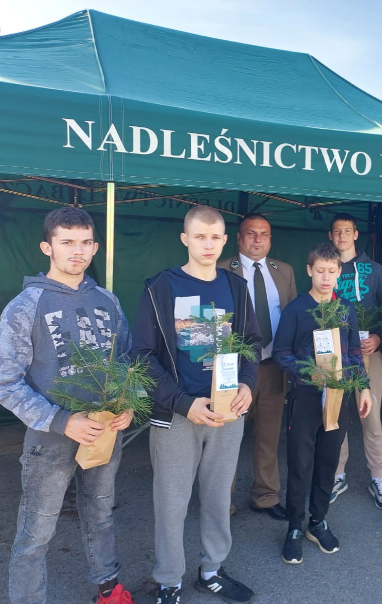 Sadzimy z Prezydentem RP | Czterech wychowanków MOW pozuje do zdjęcia z pracownikiem Nadleśnictwa ubranym w mundur. Chłopcy trzymają sadzonki drzew. W tle zielony namiot Nadleśnictwa..jpg