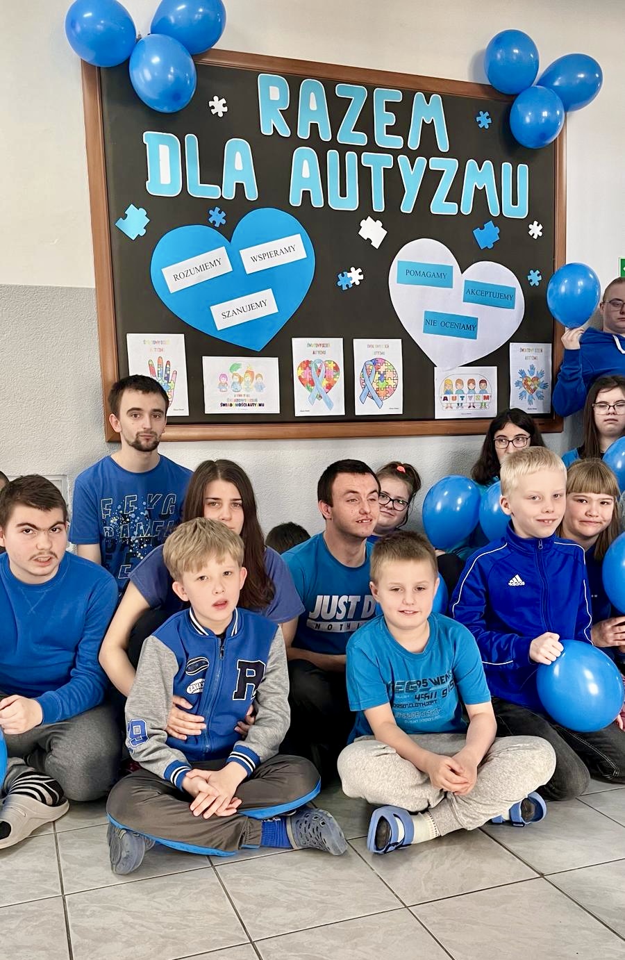 Światowy Dzień Świadomości Autyzmu | Uczniowie ubrani na niebiesko siedzą pod tablica na temat autyzmu.jpg