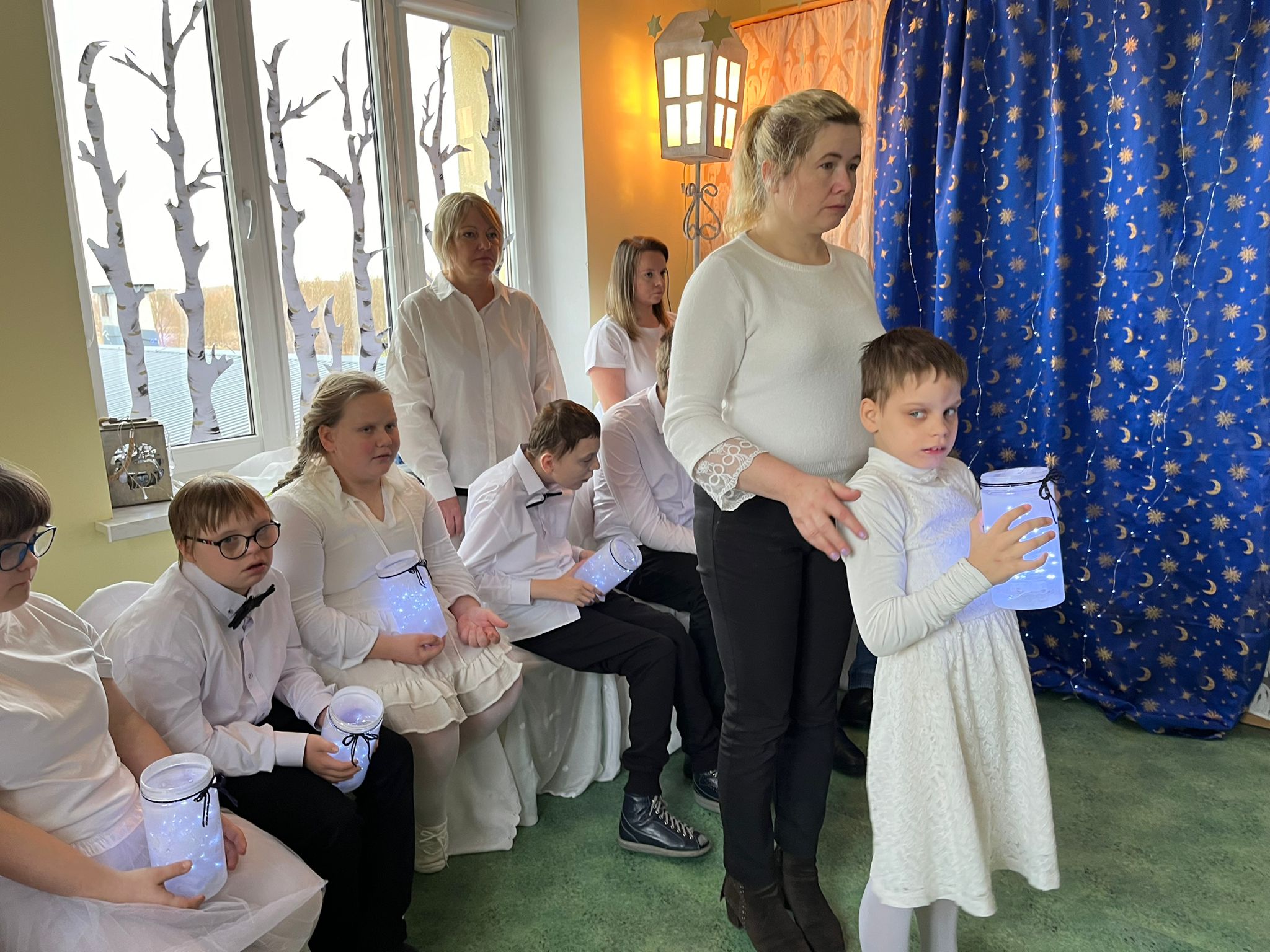 Taniec NOEL | Dziewczynka z nauczycielem stoi n środku sali , w tle siedzą pozostali uczniowie.jpg