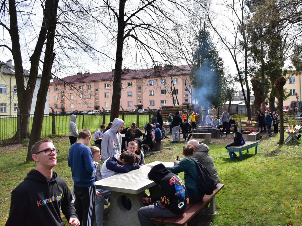 Czekając na Święta | Grupa chłopców w plenerze siedząca na ławeczkach, przy stolikach lub stojąca pomiędzy. Z tyłu palący się grill, drzewa.JPG