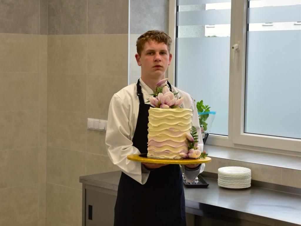 Goście w Zespole Placówek | Chłopiec w białej koszuli i czarnym fartuchu stoi wewnątrz pracowni kulinarnej trzymając w dłoniach tacę z wysokim, jasnym, udekorowanym kwiatam.JPG