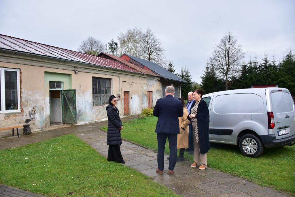 Goście w Zespole Placówek | Grupa dorosłych stoi przed wejściem do niskiego budynku, rozmawiając ze sobą. Za nimi jasny samochód dostawczy_.JPG