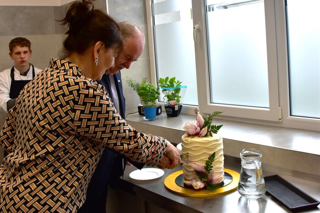 Goście w Zespole Placówek | Kobieta i mężczyzna wspólnie kroją wysoki, udekorowany kwiatami tort stojący na blacie. Za nimi widoczny chłopiec w fartuchu i białej koszuli_.JPG