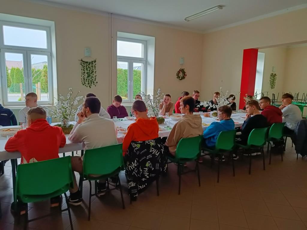 Wielkanoc w Ośrodku | Chłopcy w kolorowych ubraniach siedzą dokoła długiego stołu nakrytego białym obrusem. Na stole wielkanocne stroiki, talerze z potrawami_.jpg