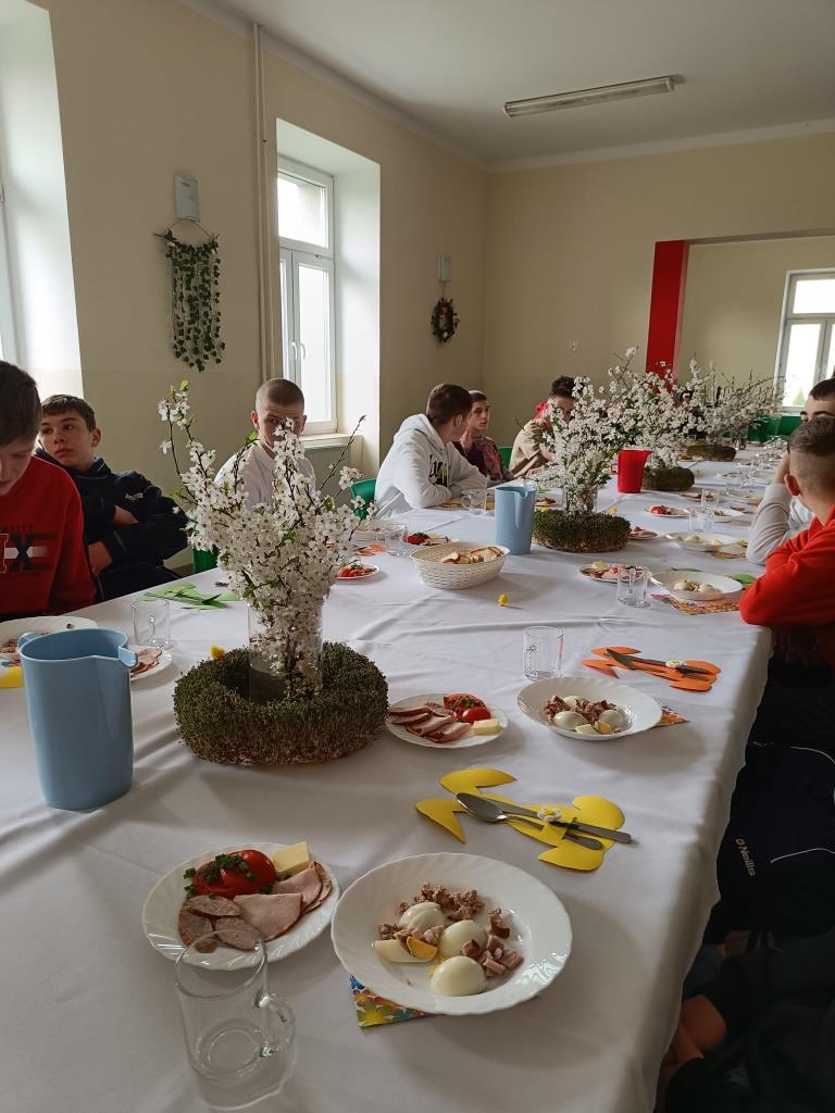Wielkanoc w Ośrodku | Stół nakryty do wielkanocnego śniadania, talerze z jajkiem, szynką, sałatką, pośrodku stroiki. Za stołem siedzi kilku chłopców_.jpg