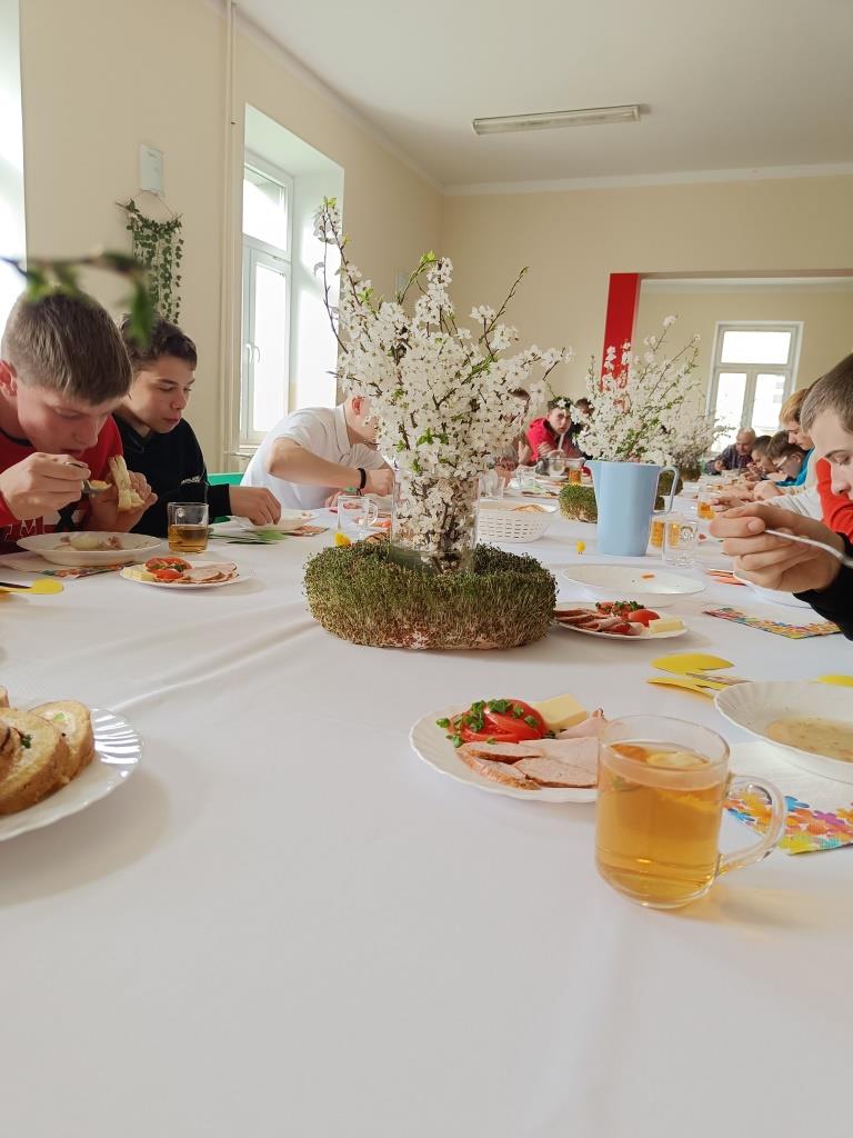 Wielkanoc w Ośrodku | Zdjęcie z perspektywy blatu stołu, na stole szklanka z herbatą, talerze z jedzeniem, stroik. Za stołem kilku chłopców w trakcie jedzenia_.jpg