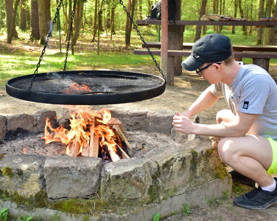 Na rowerach po okolicy | Chłopiec w czapeczce z daszkiem kuca przy cembrowinie z palącym się ogniskiem. Nad ogniem ruszt podwieszanego grilla. Z tyłu las.JPG