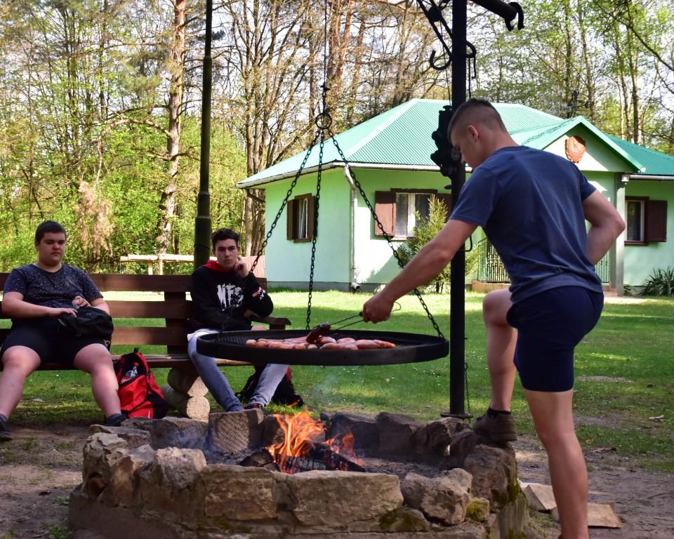 Na rowerach po okolicy | Chłopiec stoi opierając się jedną nogą o cembrowinę ogniska i przewraca piekące się na ruszcie nad ogniem kiełbaski. Dwóch innych chłopców siedzi z tyłu na ławce. W tle zielony domek.JPG