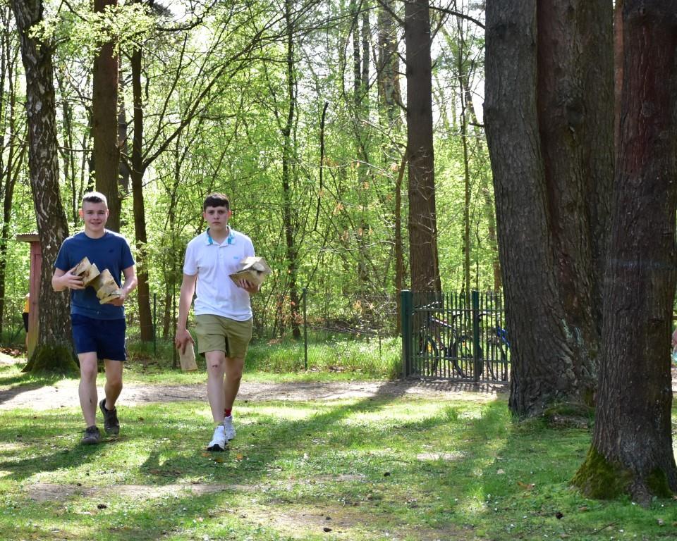 Na rowerach po okolicy | Dwóch chłopców idzie po trawie między drzewami niosąc w rękach porąbane polana.JPG