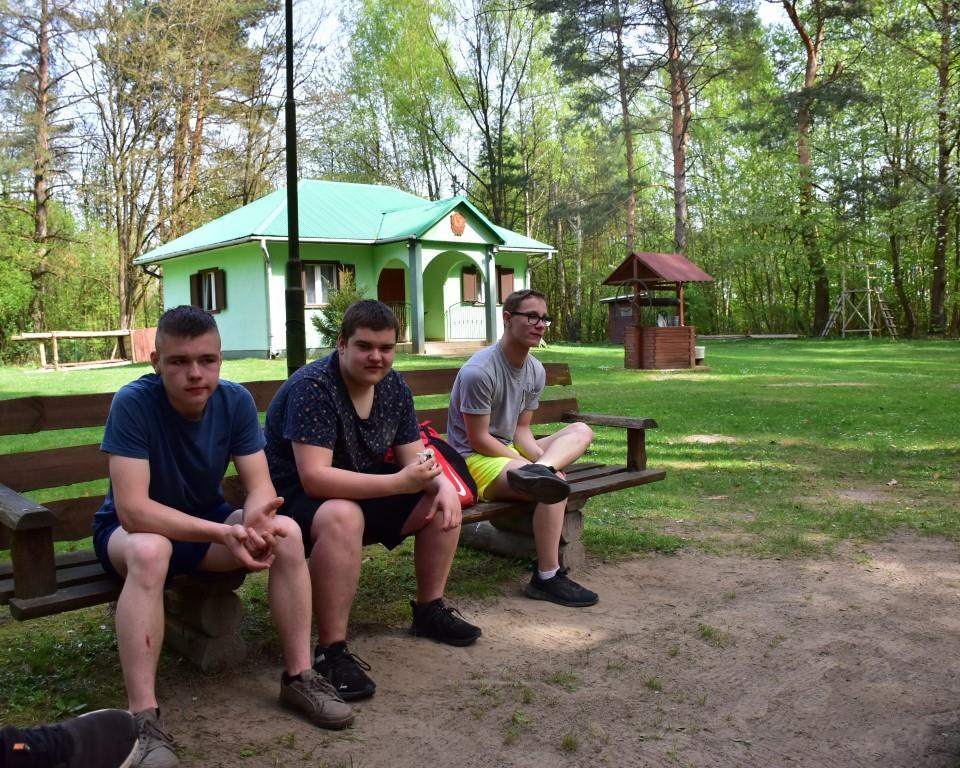 Na rowerach po okolicy | Trzech chłopców siedzi na drewnianej ławce patrząc w obiektyw. Z tyłu trawnik, na nim zielony domek, obok studnia z daszkiem. W tle las.JPG