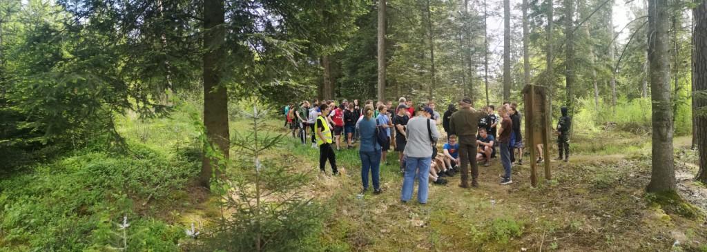 Majówka w MOW | Duża grupa osób stoi na polanie, przed nimi drewniana tablica informacyjna. Obok tablicy stoi mężczyzna w mundurze leśniczego. Zdjęcie panoramiczne..jpg