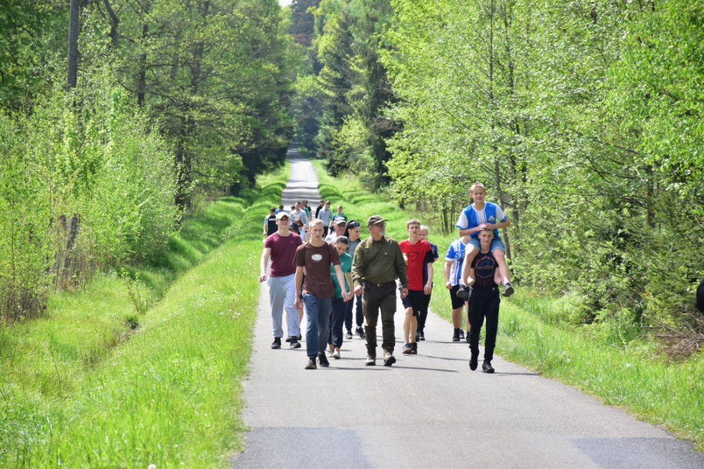 Majówka w MOW | Grupa chłoców idzie asfaltową drogą przez las, jeden z chłopców niesie drugiego na ramionach. Między chłopcami idzie mężczyzna w mundurze..JPG