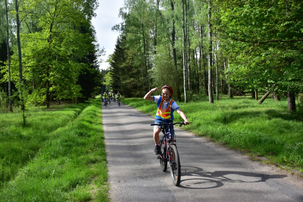 Majówka w MOW | Po asfaltowej leśnej drodze jedzie na rowerze uśniechnięty chłopiec, jedną rękę z zaciśniętą pięścią unosi w górę jak w geście zwycięstwa. W oddali inni chłopcy jadący na rowerach..JPG