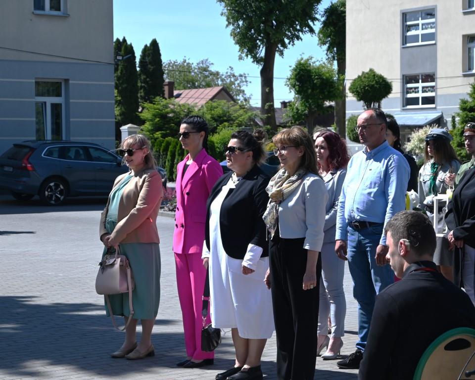 Święto Szkoły- XVII rocznica nadania imienia Jana Pawła II | Cztery odświętnie ubrane kobiety stoją w rzędzie. Za nimi mężczyzna w jasnej koszuli i kolejna kobieta, z tyłu harcerze w mundurach. W tle szarożółte budynki i zieleń.JPG