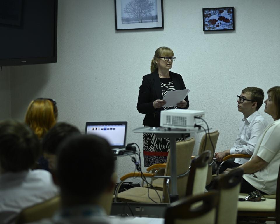 Święto Szkoły- XVII rocznica nadania imienia Jana Pawła II | Kobieta w okularach trzyma w rękach kartki papieru i wygłasza prelekcję do siedzących przed nią słuchaczy. Na środku stoi rzutnik i laptop.JPG