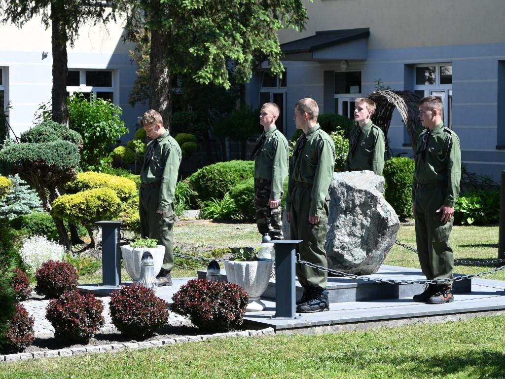 Święto Szkoły- XVII rocznica nadania imienia Jana Pawła II | Harcerze w zielonych mundurach trzymają watrę przy kamiennym obelisku. Dokoła zieleń krzewów, w tle szarożółty budynek szkoły.JPG