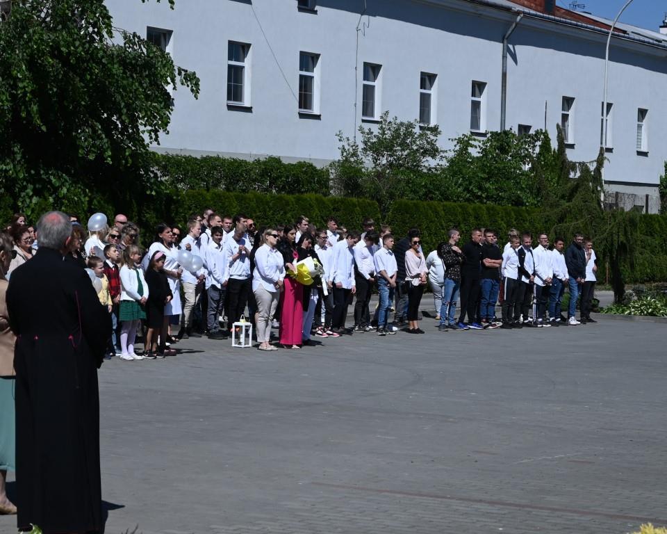 Święto Szkoły- XVII rocznica nadania imienia Jana Pawła II | Młodzież i dorośli stoją szpalerem na placu szkolnym. Przed nimi, bokiem, stoi ksiądz w sutannie. W tle żywipłot, drzewa i szaty budynek.JPG