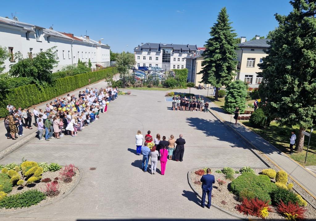 Święto Szkoły- XVII rocznica nadania imienia Jana Pawła II | Plac szkolny ze stojącymi na nim uczestnikami uroczystości i chórem. Zdjęcie z góry.jpg