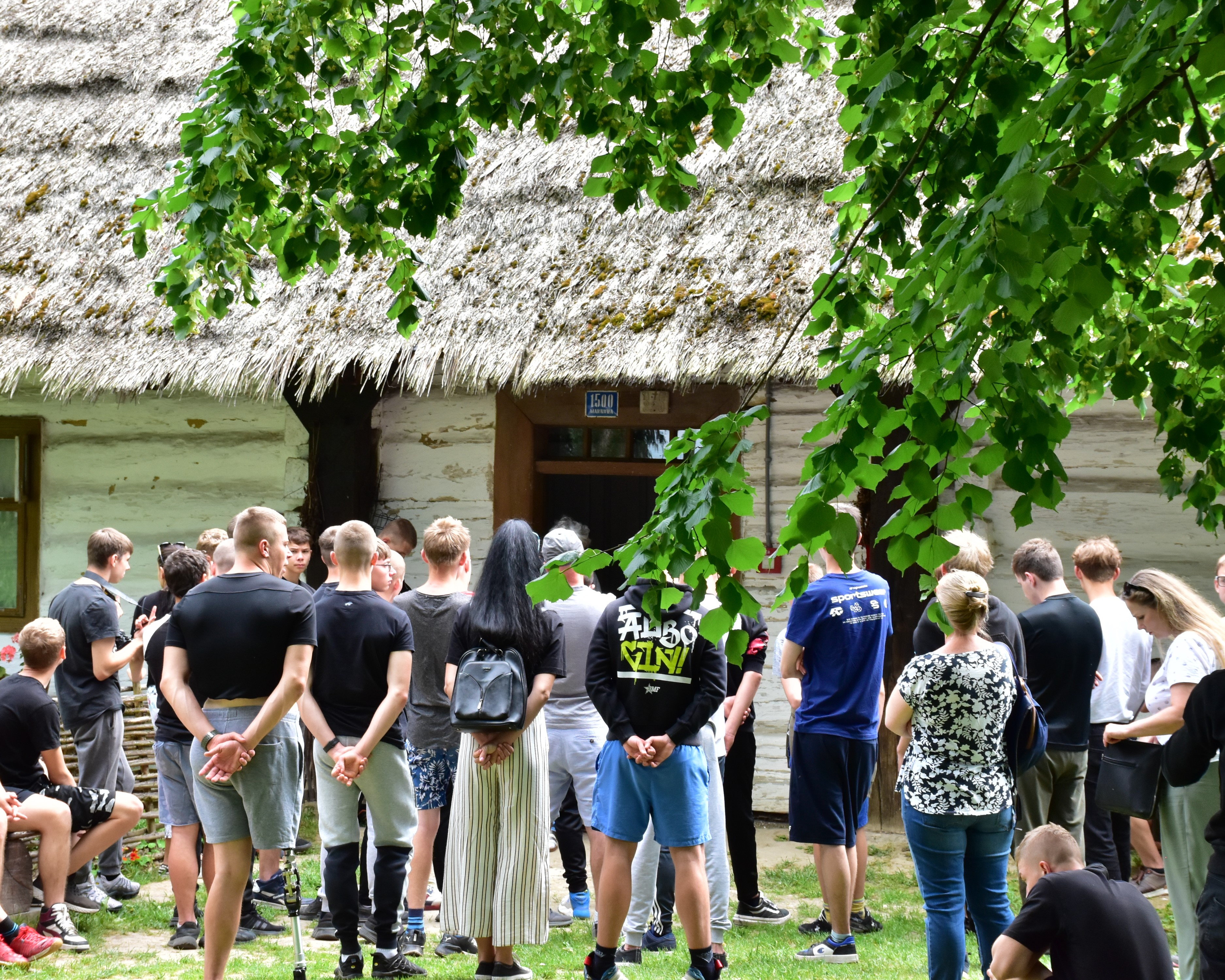 Wycieczka do Markowej | Grupa chłopców z opiekunami stoi tyłem przed chatą krytą strzechą. Z góry widoczne liście drzewa..JPG
