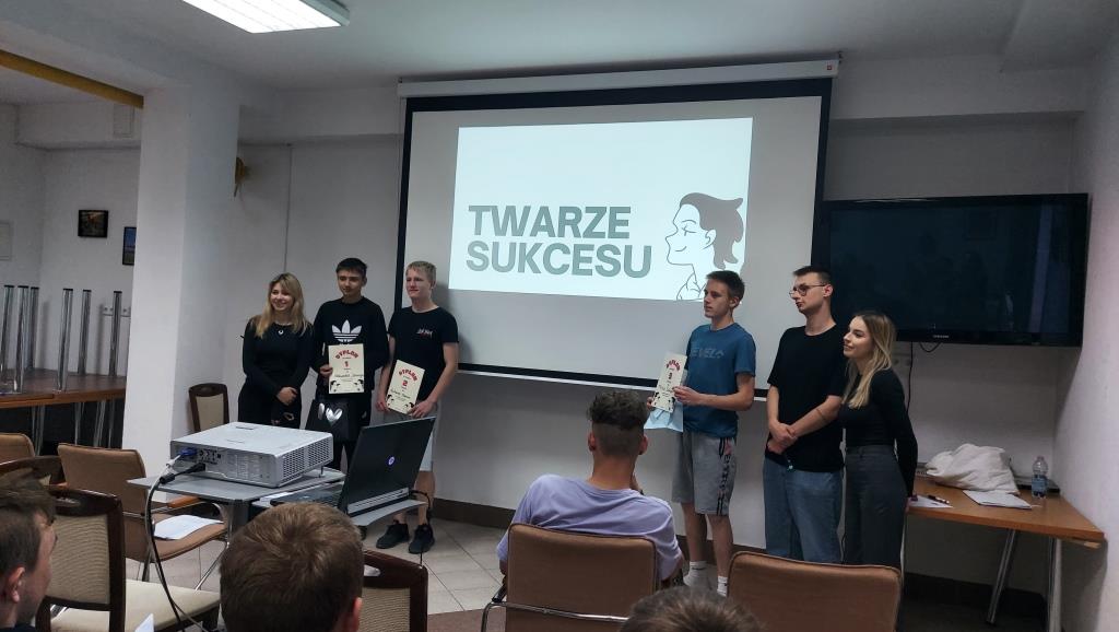 Spotkanie „Twarze sukcesu” | Przed ekranem z napisem Twarze sukcesu stoją trzej prowadzący oraz trzech chłopców z dyplomami.jpg