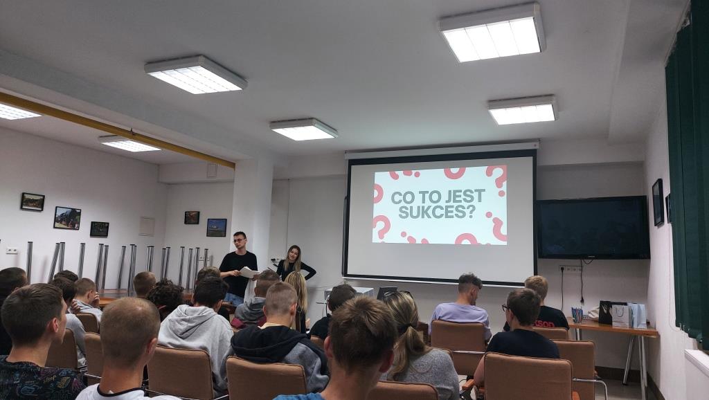 Spotkanie „Twarze sukcesu” | Zdjęcie zrobione z tyłu sali. Na krzesłach siedzi grupa chłopców, przed nimi dwoje prowadzących spotkanie i ekran z napisem Czym jest sukces.jpg