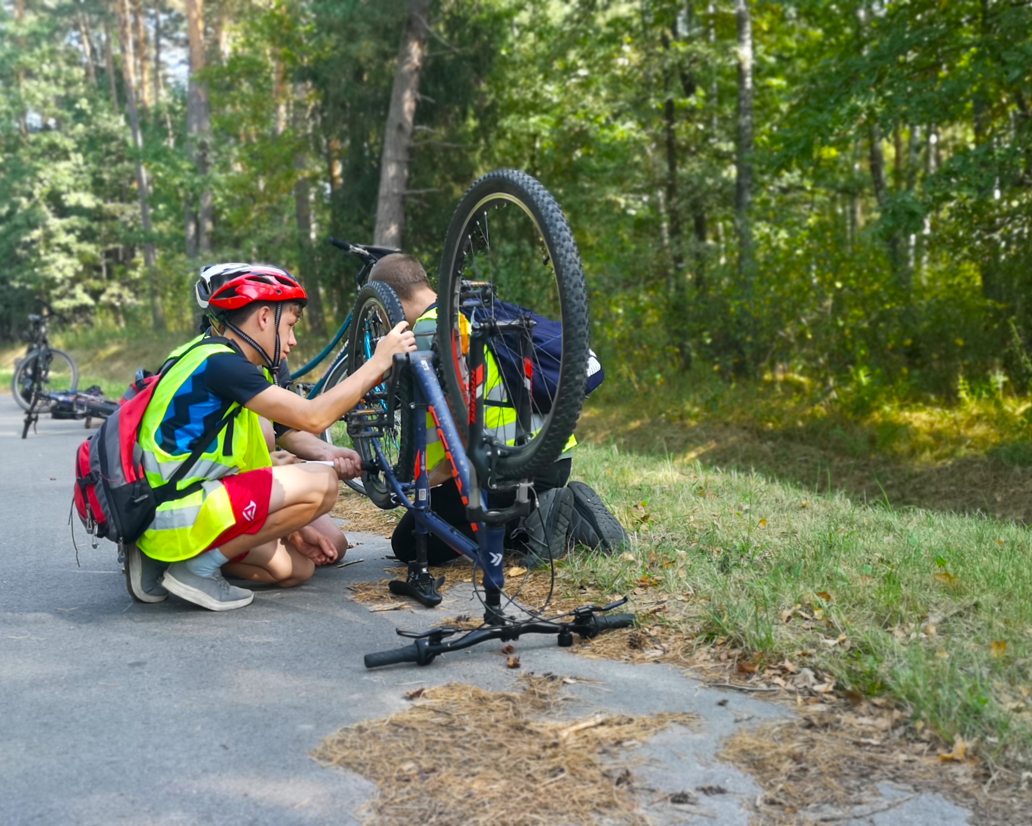 Aktywna niedziela | Na leśnej drodze stoja i leżą rowery. Przy dwóch z nich kucaja chłopcy sprawdzając stan roweru. Przy pierwszym rowerze chłopiec w kasku i kamize.jpg