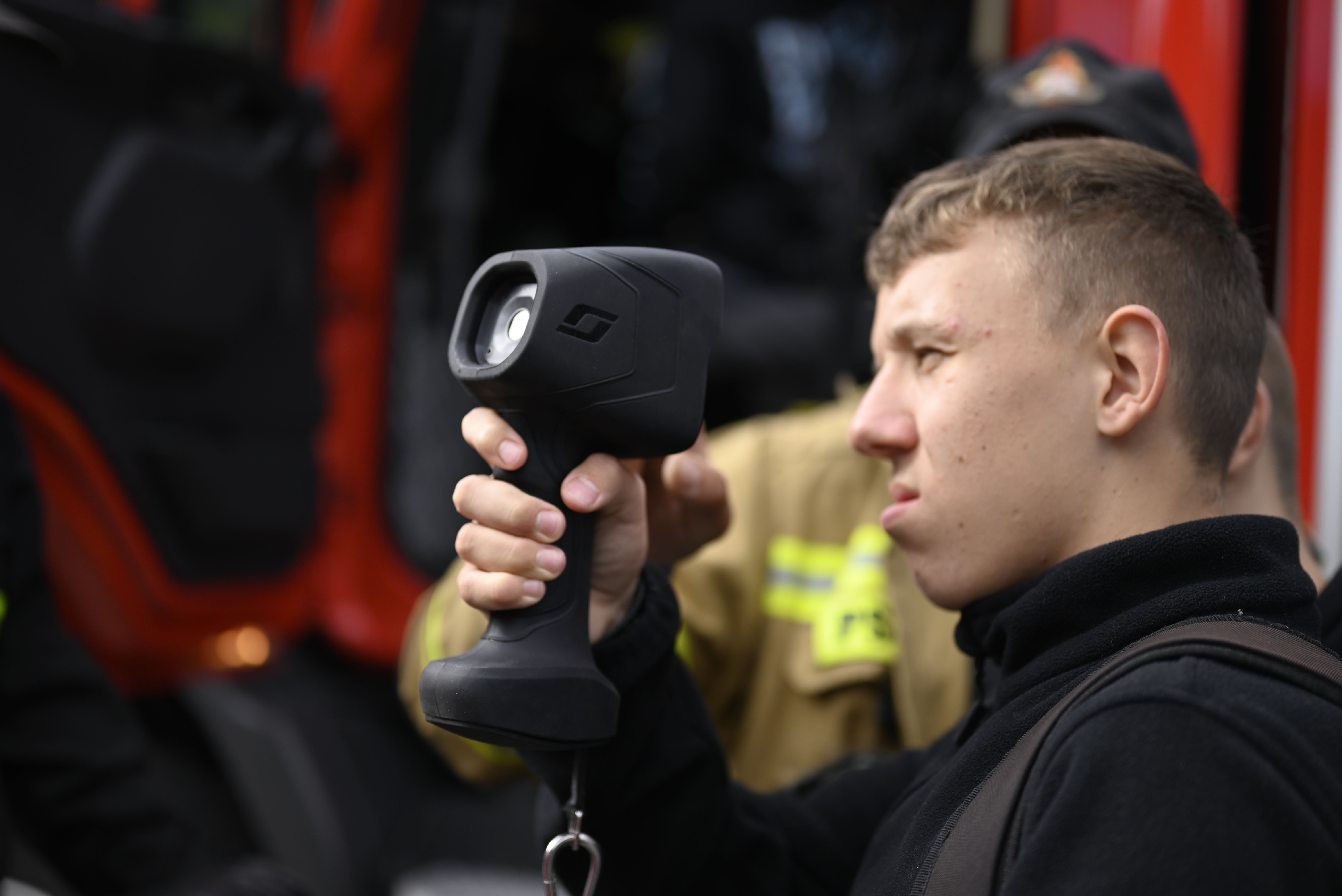 Wizyta Państwowej Straży Pożarnej | na zdjęcie na pierwszym planie nastolatek trzymający w dłoni , tyz przy oku urządzenie opzwalające parzeć w dal. Jedno oko ma zmrużone. Zdjęcie wykonane na tle samochodu strażackiego..JPG