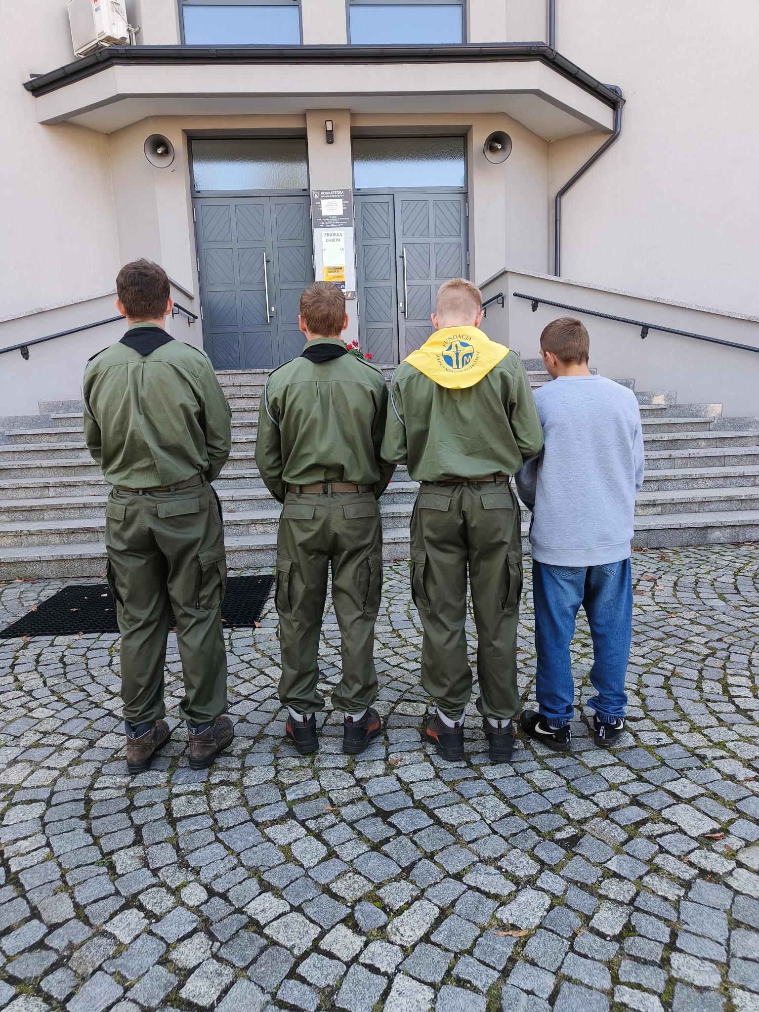 XXIV Dzień Papieski  | Przed schodami kościaoła, tyłem do obiektywu a przodem do drzwi, stoją trzej wychowankowie w mundurach harcerski obok, po prawej jeden z chłopców jest ubrany w dżnsy i bluzę..jpg