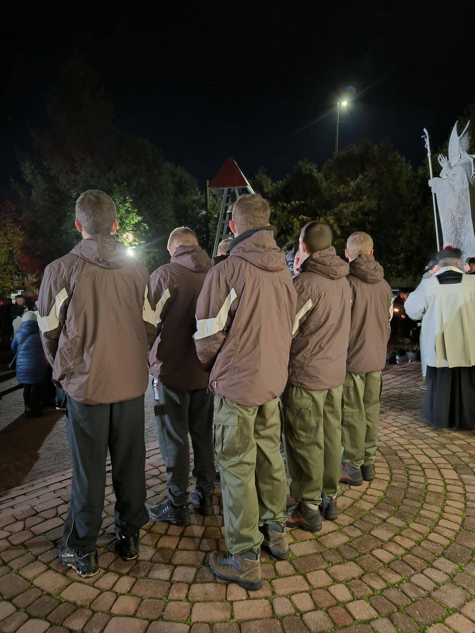 XXIV Dzień Papieski  | Grupa około dziezięciu harcerzy stoi w dwuszeregu,Stojaątyłem do obiektywu,. Po lewej stronie z tyłu stoi tyłem ksiądz ubrany w sutannę i komżę. djęcie wykonane nocą..jpg