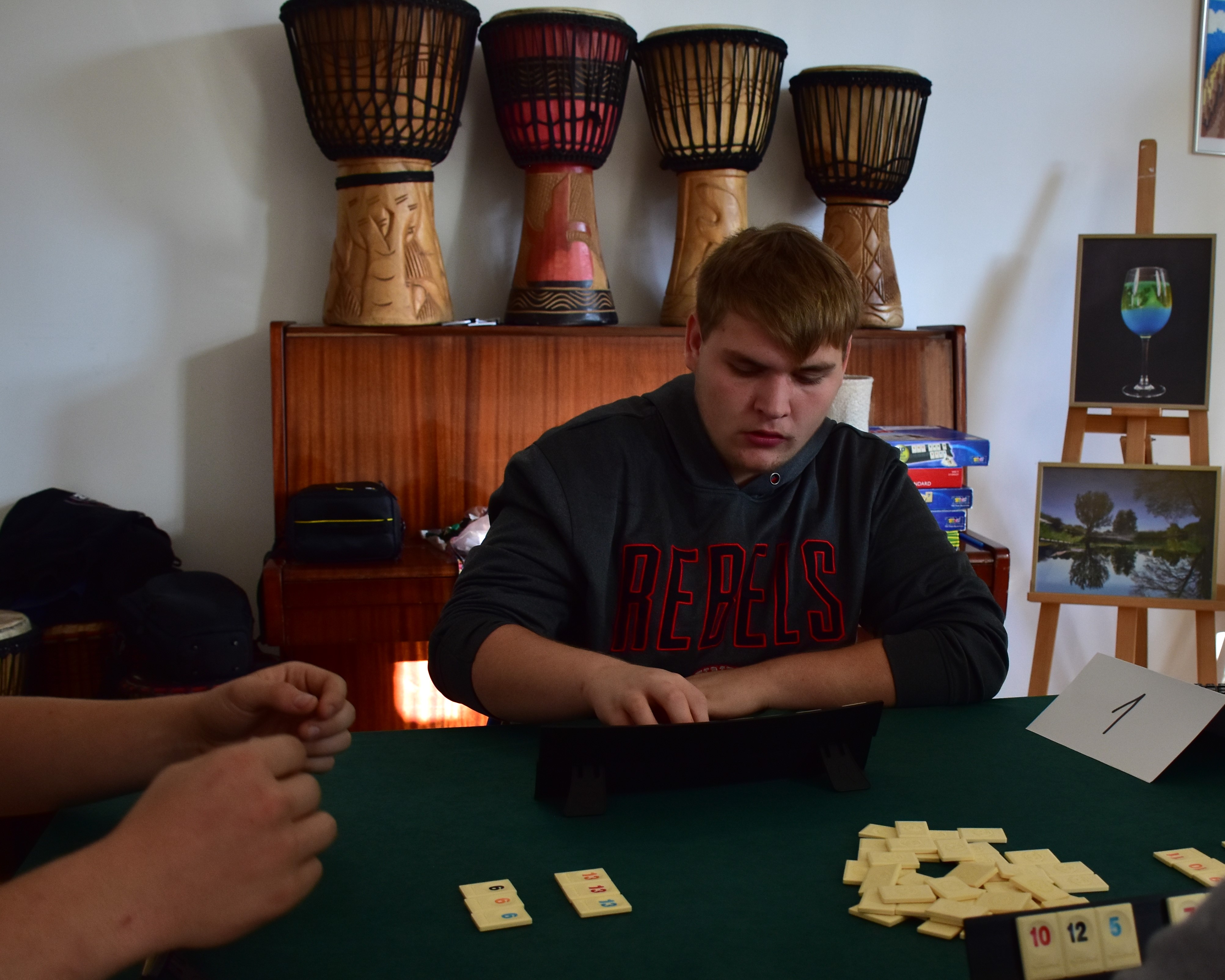 Szkolny Turniej Rummikub….. | Przy stoliku do gry  siedzi chłopiec. Trzyma w ręce kostki, a przed nim stojak. Widoczne z lewej strony ręce drugiego zawodnika.Z tyłu stoi pian.JPG