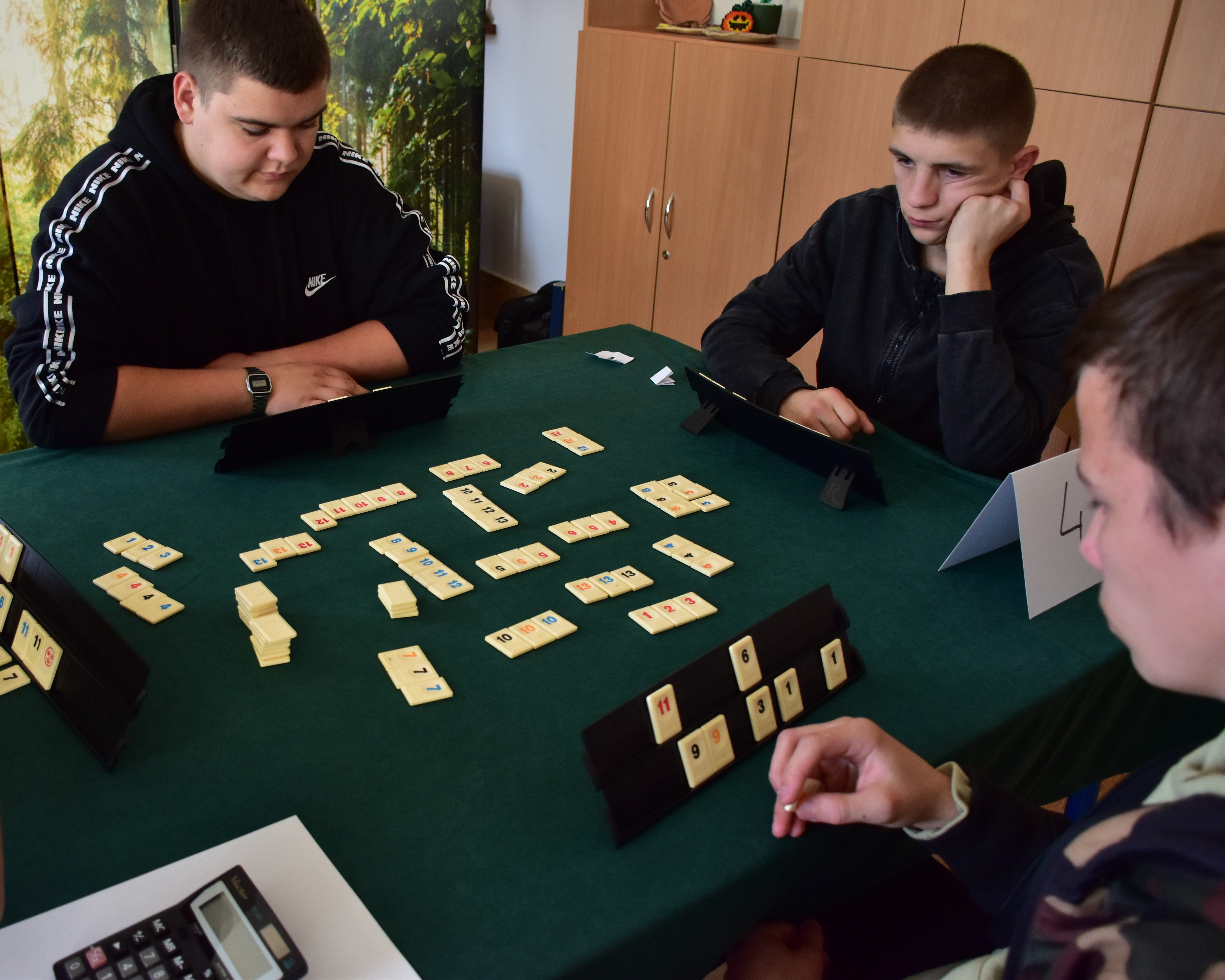 Szkolny Turniej Rummikub….. | Przy stoliku zascielonym suknem siedzi czterech zawowników. Przed każdym stoi stojak na kostki.Na stole są rozłożone kostki do gry z wyłożeń każ.JPG