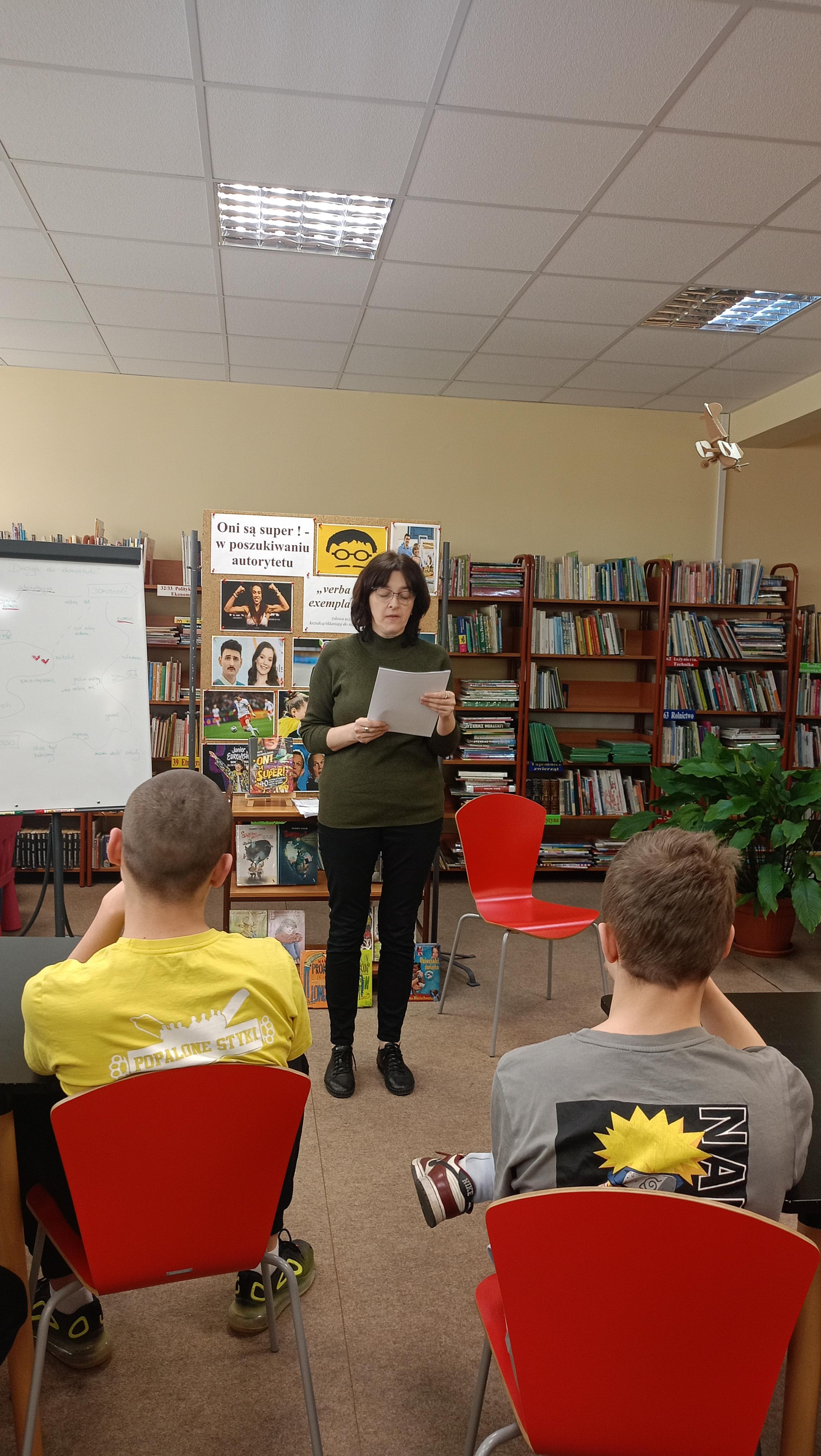LEKCJA BIBLIOTECZNA | Posród regałów z książkami siedzą wychowankowie, Przed nimi  prowadząca zajęcia, czytająca coś z katrki. Za nią tablica z hasłem spotkania Oni są super-w poszukiwaniu autorytetów oraz zdjęcia.jpeg