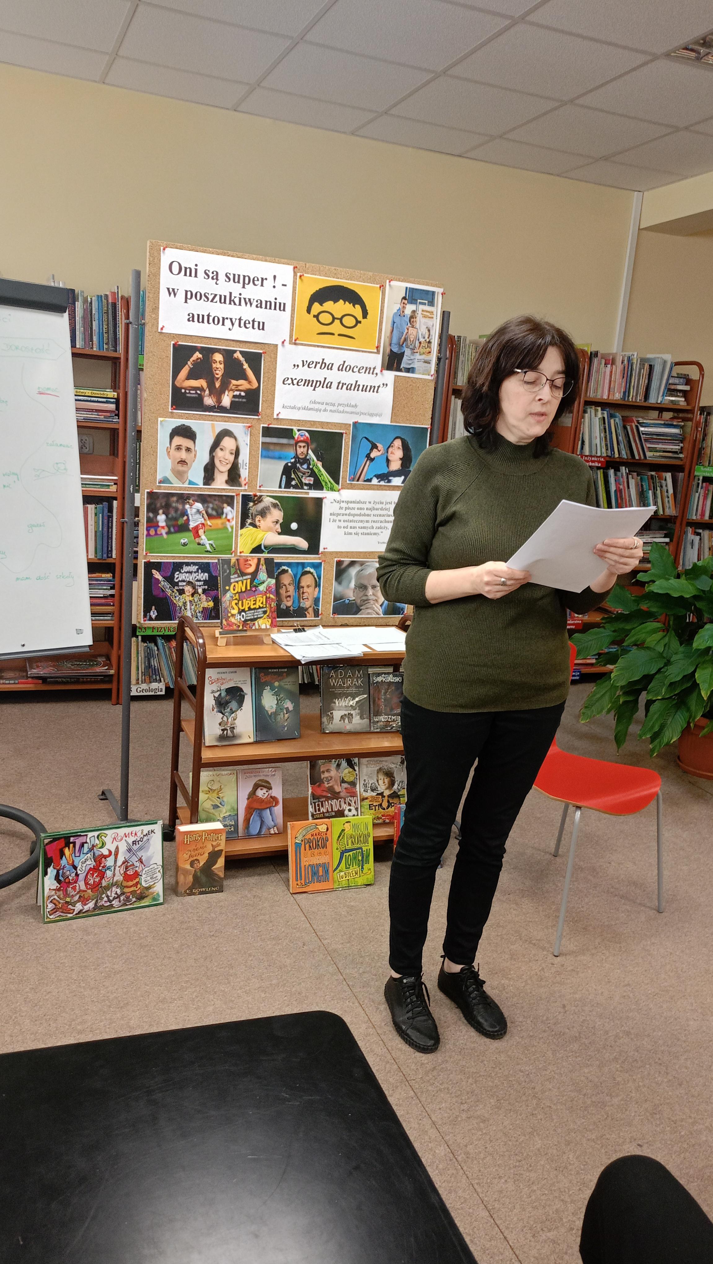 LEKCJA BIBLIOTECZNA | Zbliżenie na powadzącą zajęcia pracownikę biblioteki, która czyta coś z kartki. Za nia regały i półki z książkami oraz tablica z hasłem spotkania.jpeg