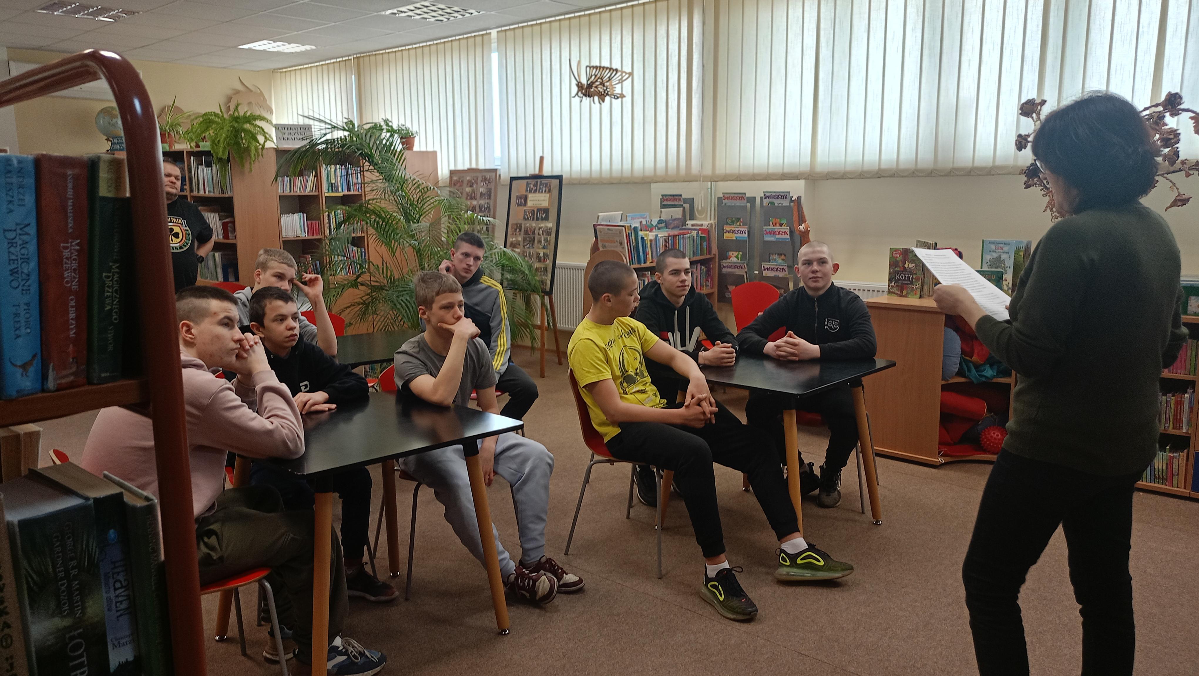 LEKCJA BIBLIOTECZNA | Zdjęcie wykonane w sali ibibliotecznej, w której wychowankowie siedzą przy dwóch stolikach trarzami zwróceni ku prowadzącej zajęcia pracownicy biblioteki..jpeg