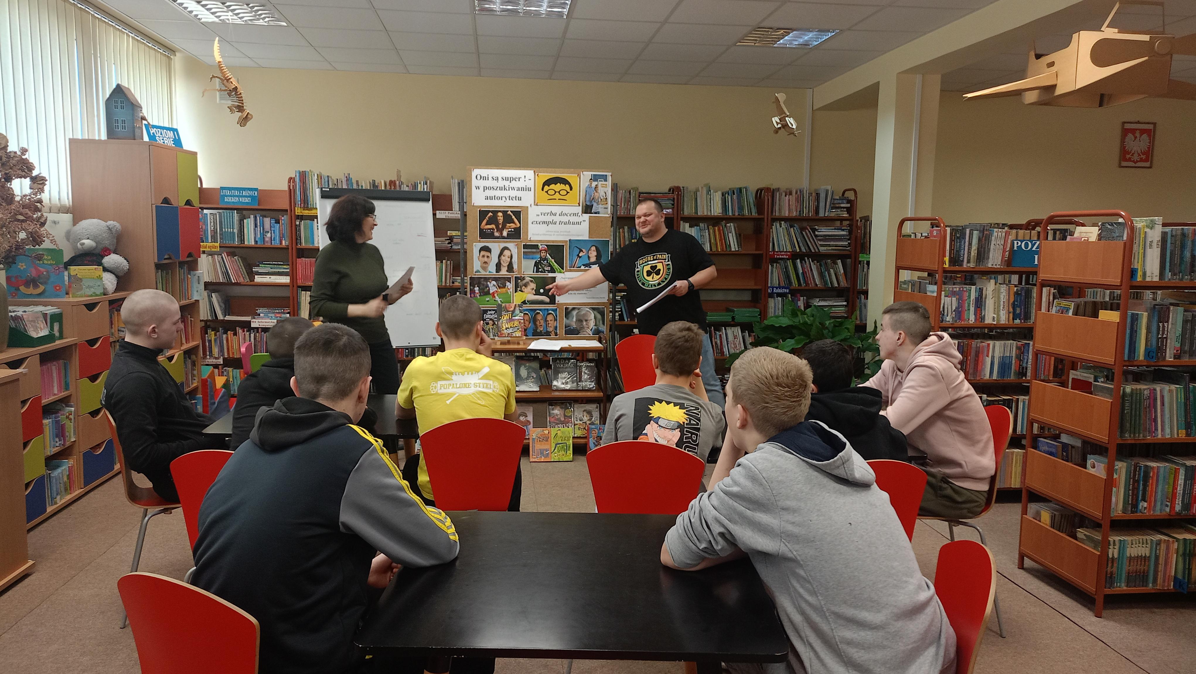 LEKCJA BIBLIOTECZNA | W sali bbiblotecznej wychowankowie siedzą przy stolokach, a przed nimi prowadzący mężczyzna i kobieta gestykulują i śmieją się. Za nimi tablica z hasłem spotkania i zdjęciami..jpeg