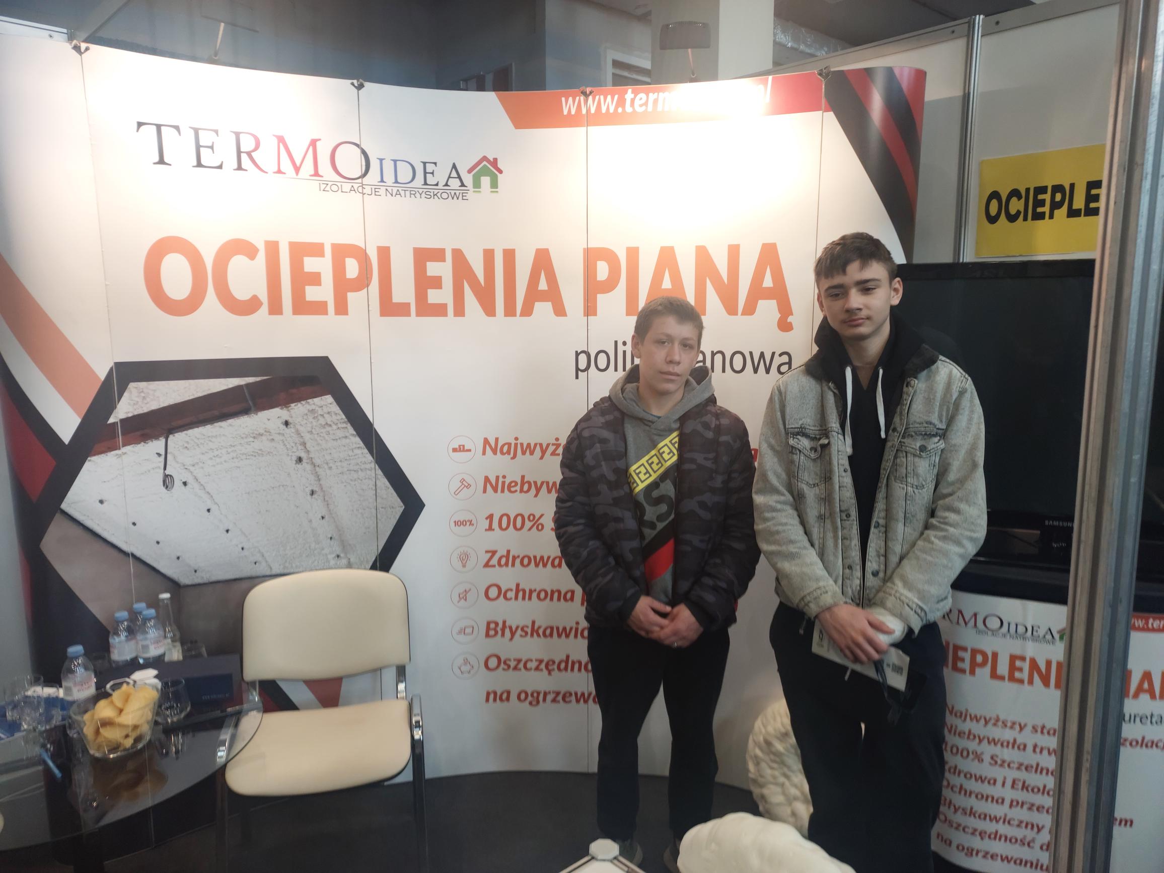 Połączenie przyjemnego z pożytecznym  | Dwaj wychowankowie stoją w boksie z napisem ocieplenie. Za nimi ogromny banner z napisem oceplanie piianka pur i wyliczonymi jej korzyściami..jpg