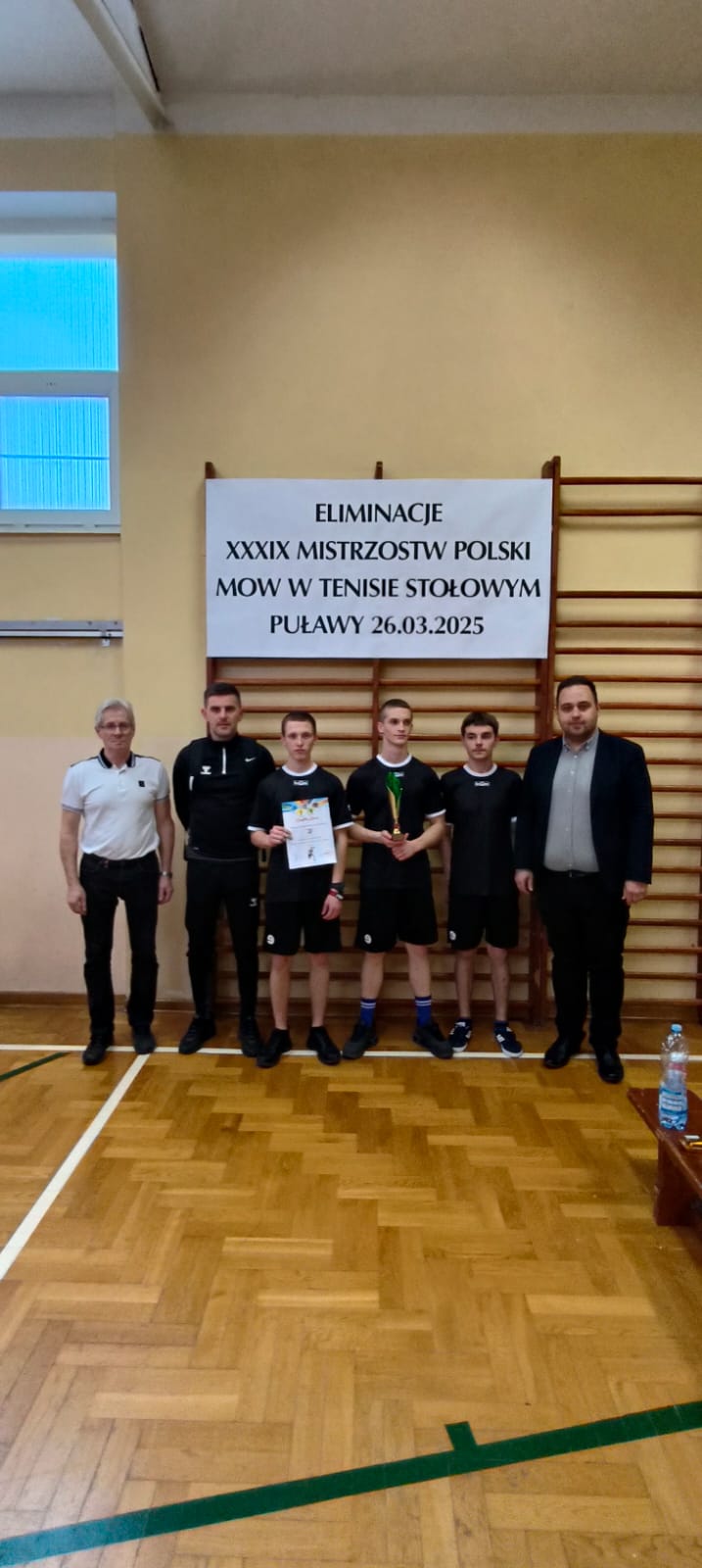 Turniej Eliminacyjny XXXIX Mistrzostw Polski MOW w Tenisie Stołowym  | Pod tablicą z nazwą Eliminacje XXXIX Mistrzostw Polski w Tenisie stołowym. Puławy 26.03.2025r. stoją trzej nasi zawodnicy wraz z trenerem oraz dwoma mężczyznami..jpg