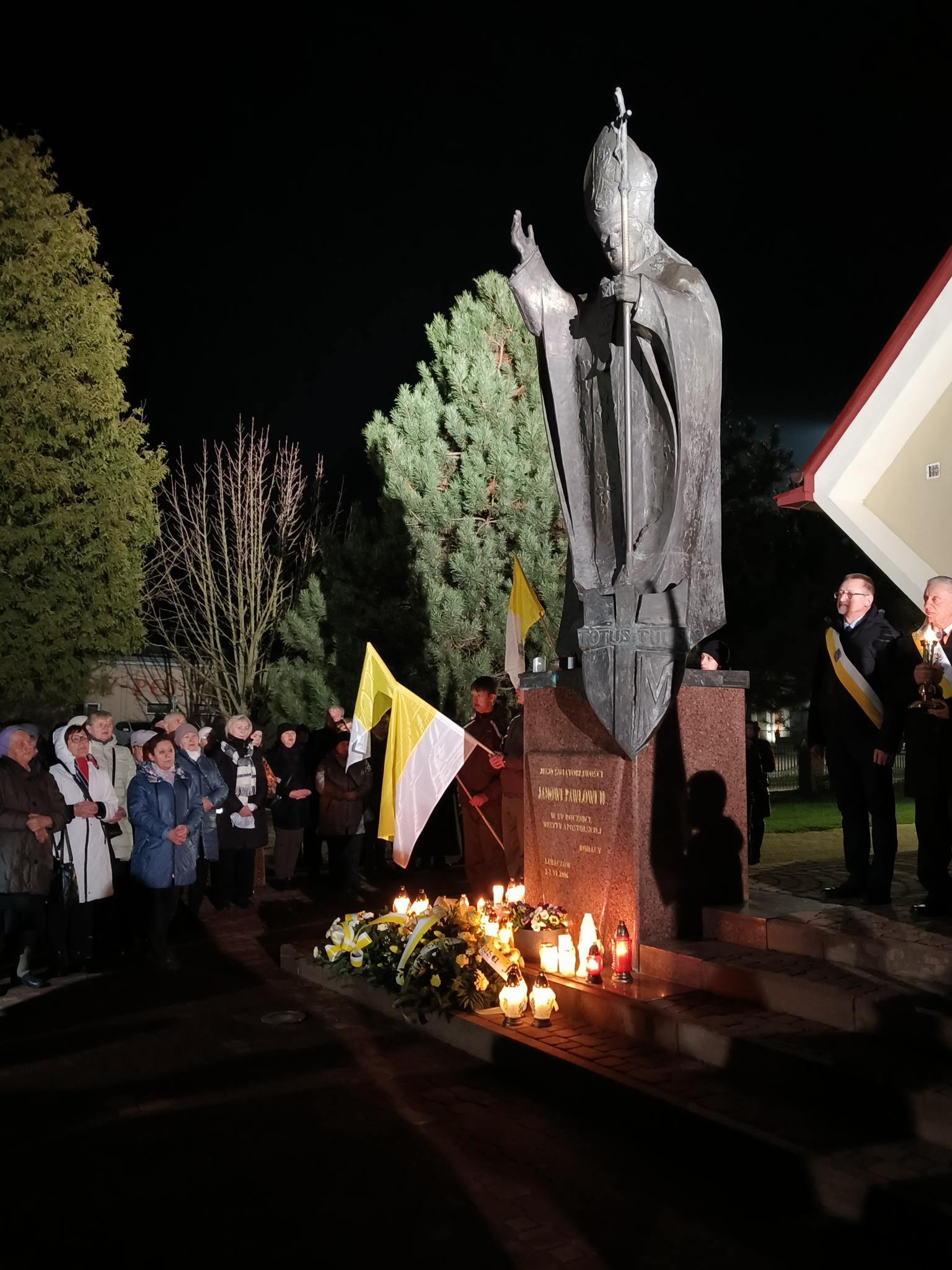 XX Rocznica śmierci św. Jana Pawła II | Zbliżenie na pomnik Św. Jana Pawła II przed krórym zostały złożone wiązanki i zapalone znicze. Za pomnikiem poczet sztandarowy, obok zgromadzeni wierni..jpg