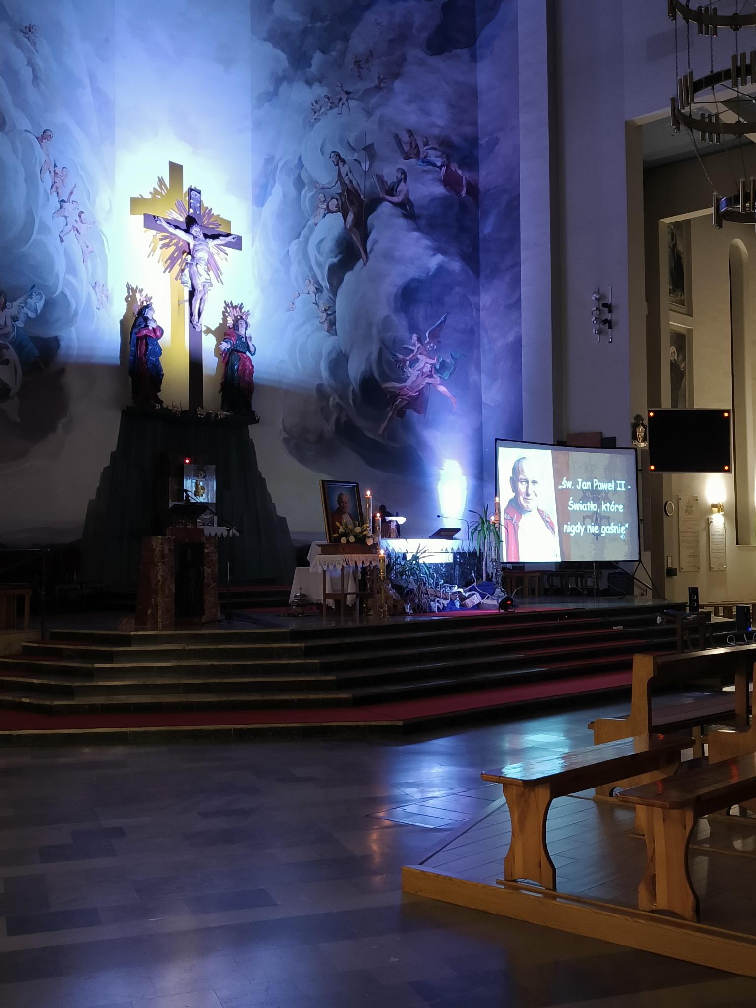 XX Rocznica śmierci św. Jana Pawła II | Ujęcie ołtarza w Konkatedrze. W półmroku, podświetlony tylko krzyż w ołtarzu . Z boku tellebim na któym wyświetlony jest sajd z napisem Św Jan Paweł II światło, ktre nigdy nie gaśnie..jpg