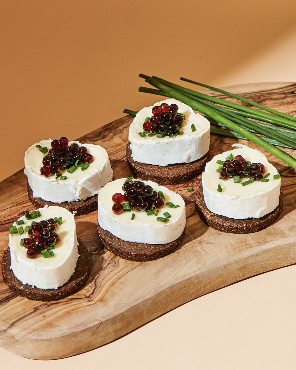 Ziegenkäse-Häppchen auf Pumpernickel mit Essigperlen und Schnittlauch auf einem Holzbrett angerichtet.