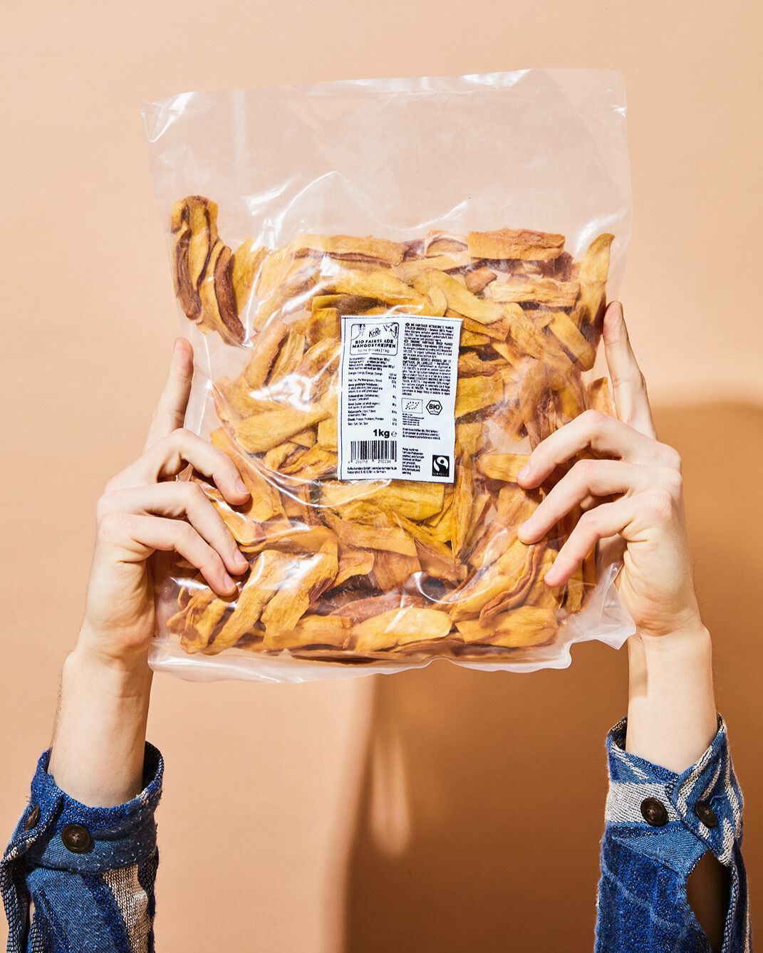 Zwei Hände halten eine transparente Plastikverpackung mit 1 kg getrockneten Mangostreifen von KoRo in die Höhe. Die Mango ist goldgelb und leicht gebräunt. Auf der Verpackung ist ein weißes Etikett mit Produktinformationen und dem Fairtrade-Siegel zu erkennen. Der Hintergrund ist ein schlichter, pfirsichfarbener Ton.