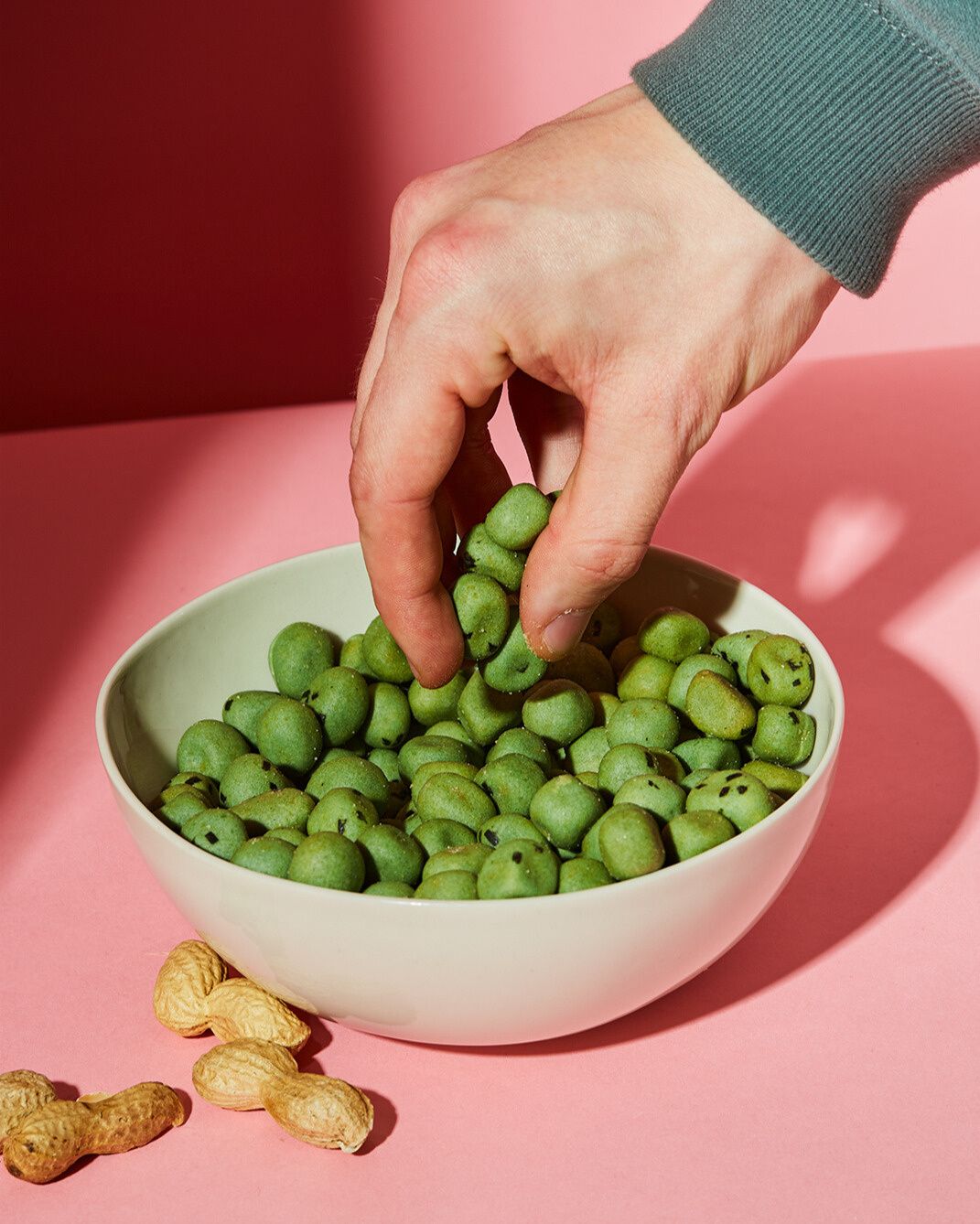 Eine Hand greift in eine weiße Schale voller hellgrüner, dragierter Erdnüsse, die auf einem rosa Hintergrund steht. Neben der Schale liegen einige Erdnüsse in ihrer natürlichen Schale.
