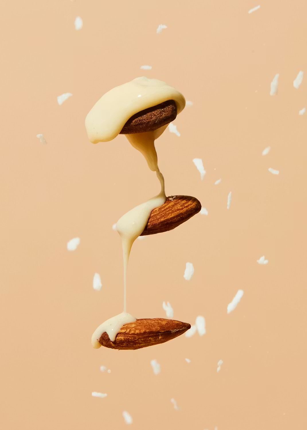 Drei vertikal schwebende Mandeln, über die eine cremige weiße Glasur fließt, umgeben von kleinen weißen Splittern vor einem pfirsichfarbenen Hintergrund.