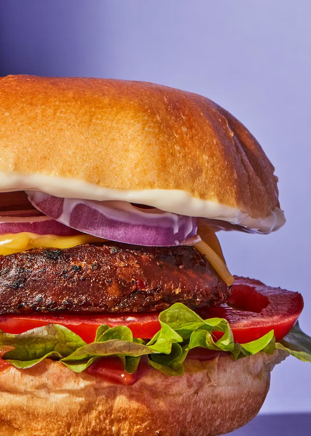 Ein saftig belegter Burger mit frischem Salat, Tomaten, roten Zwiebeln und cremiger Sauce vor einem violetten Hintergrund.